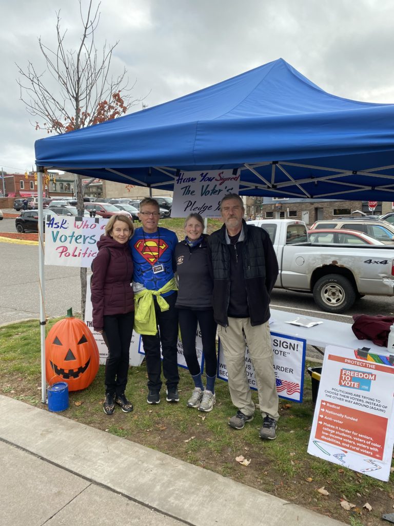 (Image Description: Bob (superman shirt) and a group of three of his core volunteers at the Marquette Farmers Market in October. Behind them is their canopy, table, a series of pro-voter signs hung from the table and canopy, as well as a plastic Halloween pumpkin.)