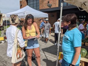 A photo of 3 women in a farmer's market. One women wears a teal shirt that says "Voters Not Politicians" on the front. The other two women stand facing her, filling out a petition on a clipboard.