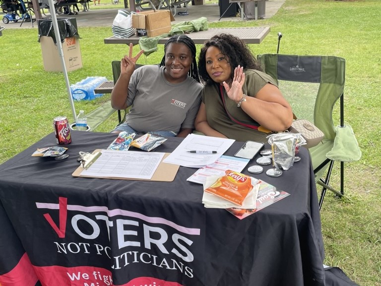 Two people seated at a table under a canopy at a park with a "Voters Not Politicians" tablecloth, holding up peace and wave gestures.  Text Tablecloth: "VOTERS NOT POLITICIANS We fight and win for Michigan voters"