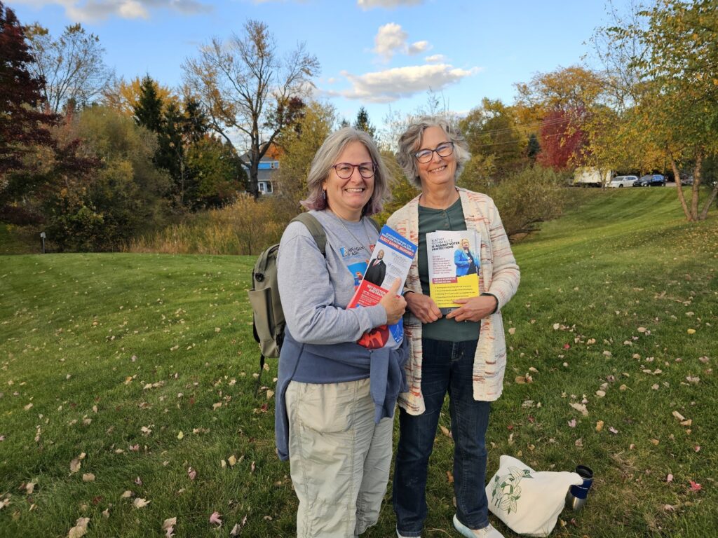 Two women stand on a grassy lawn with autumn trees in the background, holding flyers and smiling at the camera.