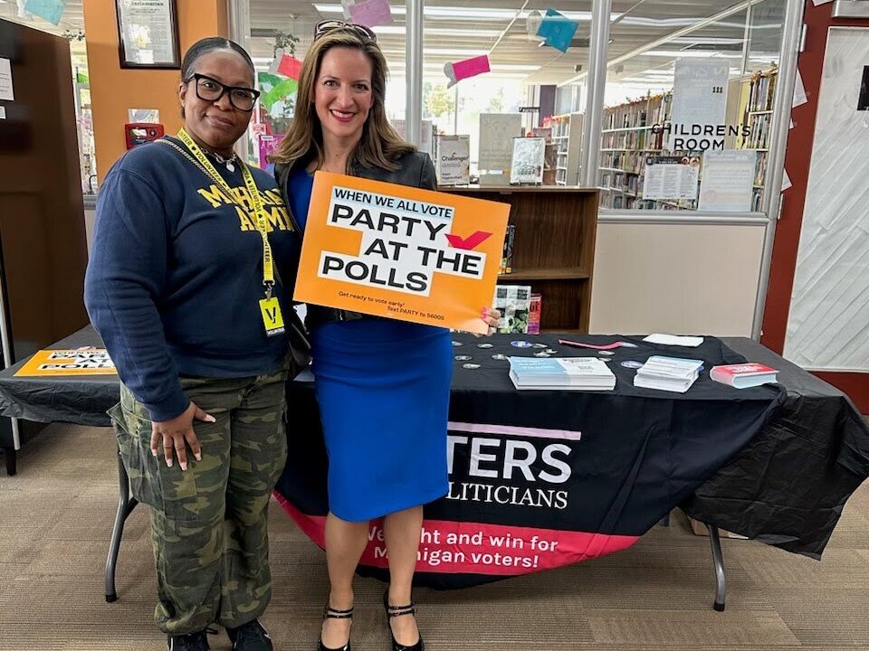 Two people in a library, one (Secretary of State Jocelyn Benson) holding a "Party at the Polls" sign. They are both standing in front of a table with a Voters Not Politicians tablecloth