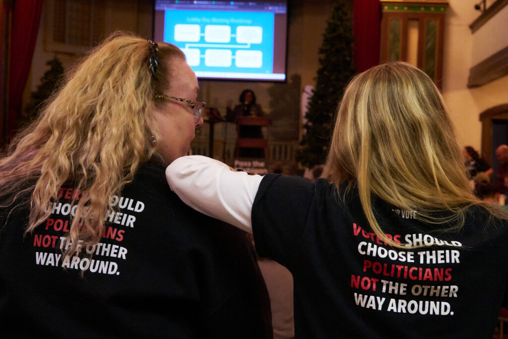 Two women in a meeting hall wearing shirts with political messages on their backs. Transcribed Text: "VOTERS SHOULD CHOOSE THEIR POLITICIANS NOT THE OTHER WAY AROUND."