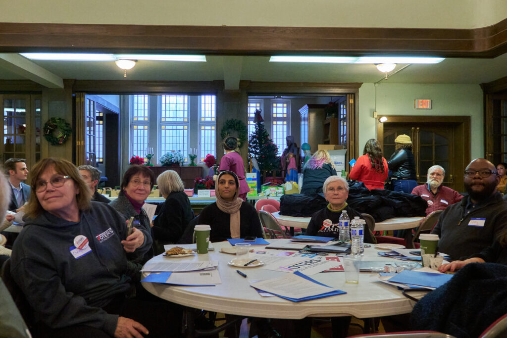 Five people sit around a table, facing the camera but looking past, towards a presentation or speaker. More people at tables are visible in background. A sign on the table reads "Voters Not Politicians"