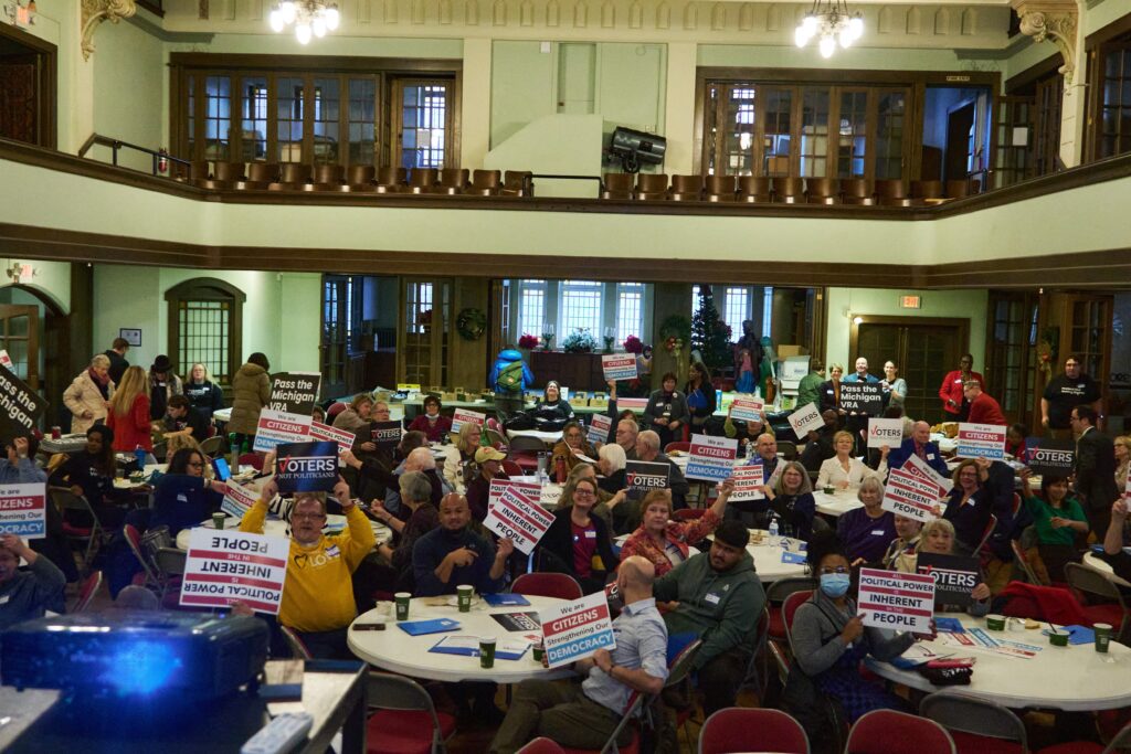 A diverse crowd of people sits around tables in a spacious room, holding various signs that read "All political power is inherent in the people" and "Voters Not Politicians"