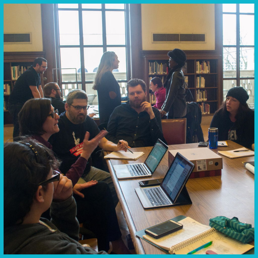 People sitting and standing around a table with laptops in a library setting.