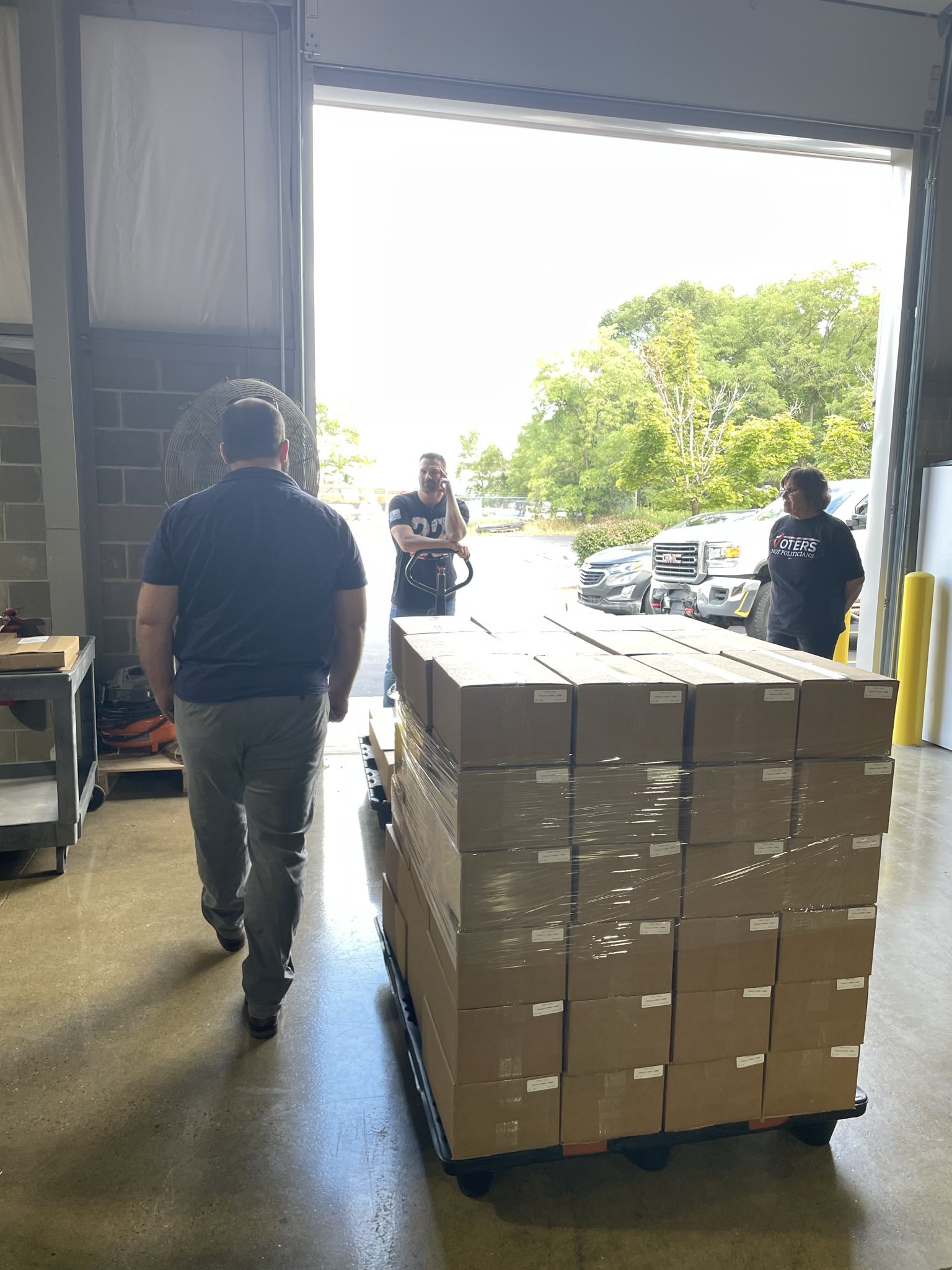 Warehouse interior with pallet of boxes and three people, one pushing the pallet. Transcribed Text: The shirt of the person standing outside to the right reads "VOTERS."