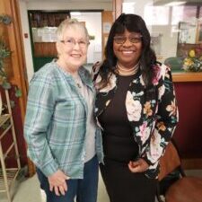 Two smiling women standing side by side indoors, one wearing a plaid shirt, the other a floral jacket.