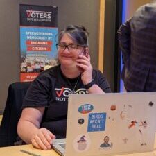 Woman smiling while talking on the phone and using a laptop covered in stickers. Banner in the background reads "VOTERS NOT POLITICIANS."